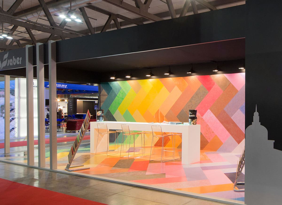 L'immagine raffigura un caso reale di allestimento fieristico: l'allestimento dello stand Saber Color realizzato per Made Expo di Milano. In particolare, nell'imagine si vedono due inquadrature. La prima a sinistra mostra un particolare dell'ingresso; la seconda a destra mostra un particolare di un'area espositiva.