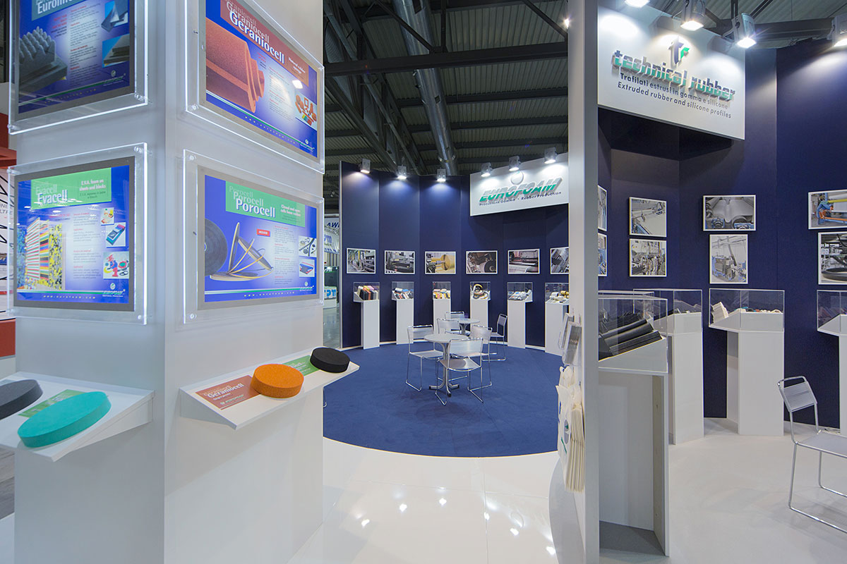 L' immagine mostra un allestimento realizzato da Diquadro per fiera Plast Milano: lo stand Eurofoma Technical Rubber. Lo stand è stato allestito creando pareti curve per poter esporre i prodotti lungo un ideale percorso che rappresenti il ciclo di lavorazione della gomma. All'interno dell' area sono stati ricavate due zone incontro con tavoli e un ufficio. I colori dello stand riprendono i colori societari delle due aziende committenti.